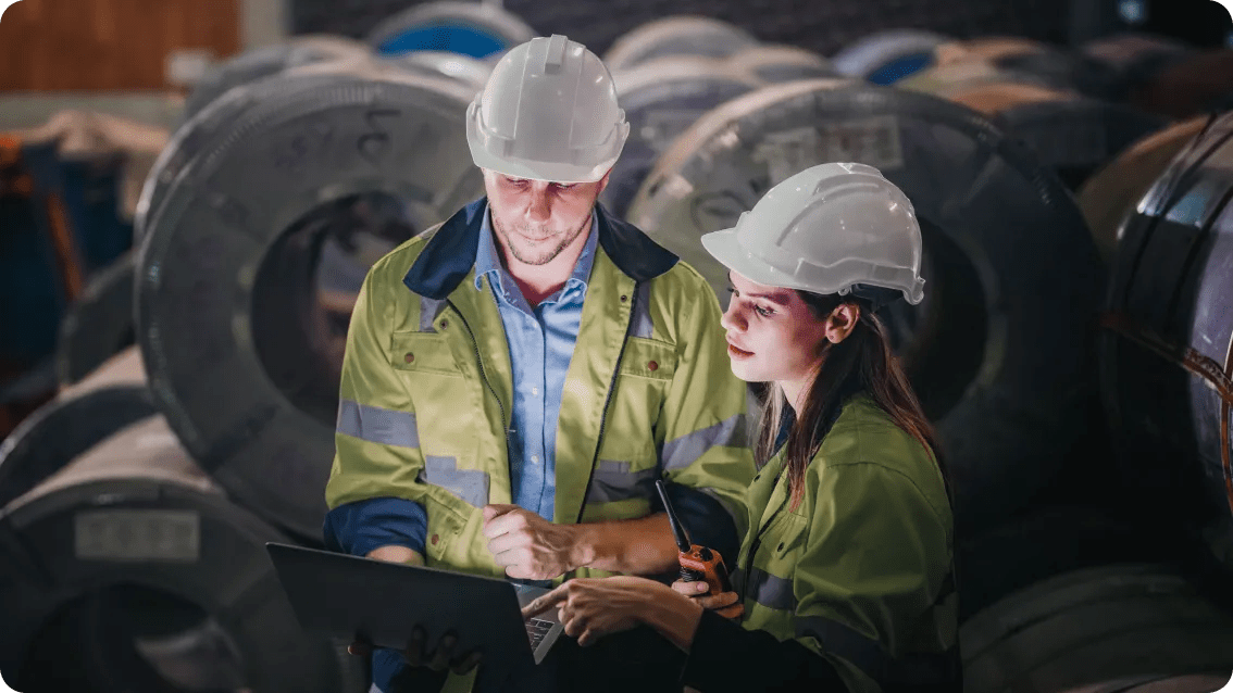 Two operators wearing hard hats, a female and a male, reviewing data on a laptop in an industrial setting.