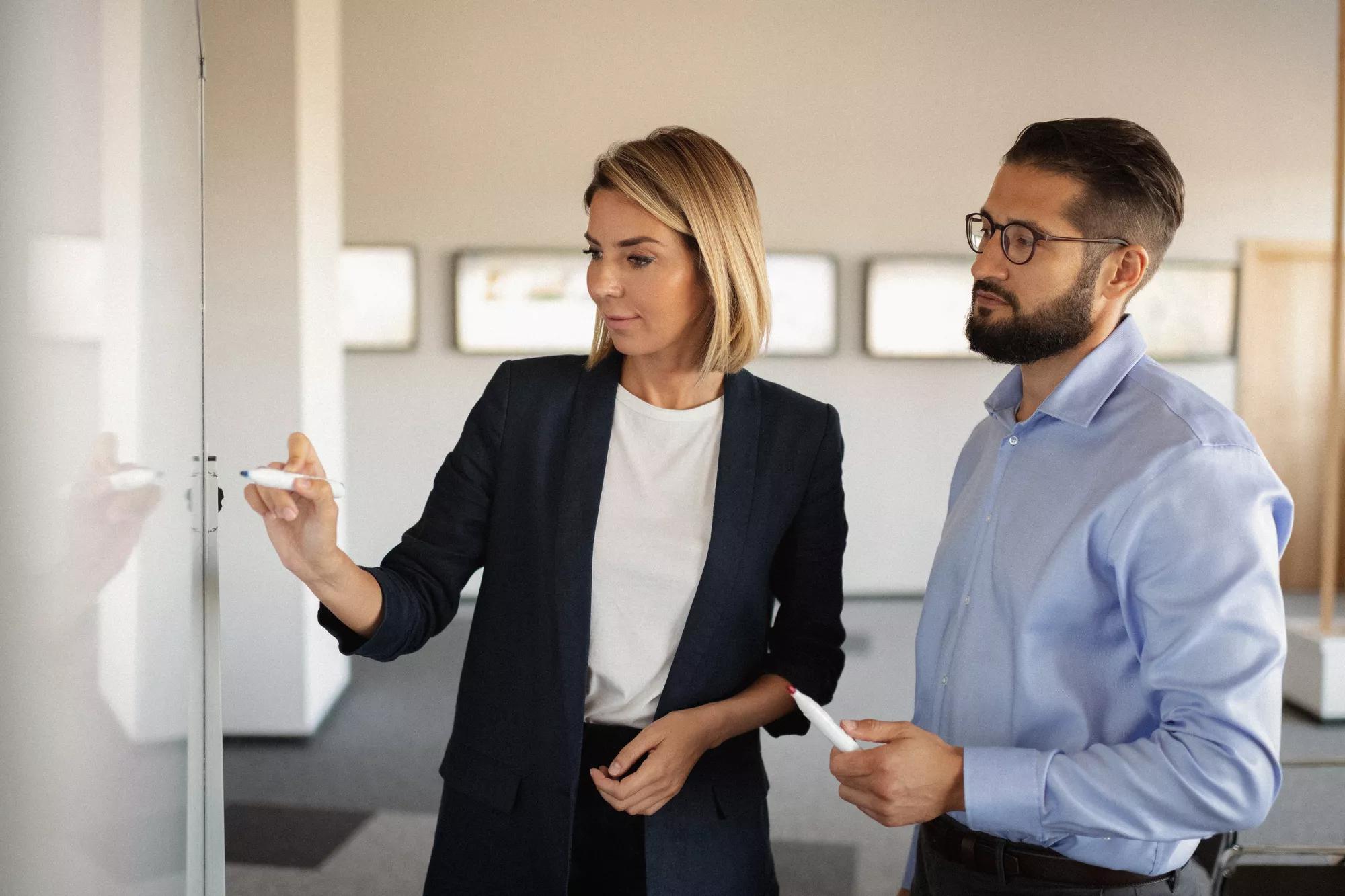 Two employees, a woman and a man, looking at and writing on a whiteboard in the office.