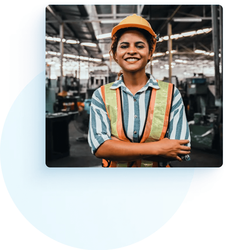 Female worker in a safety vest and hard hat standing in a warehouse in front of heavy machinery.