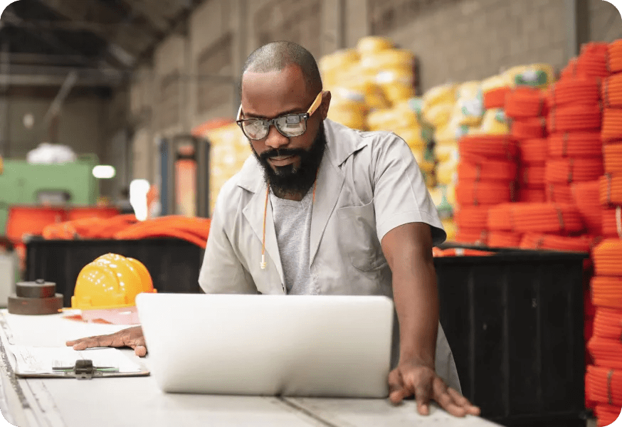 Male worker with glasses reviewing documentation and comparing it to data on a laptop on a table.